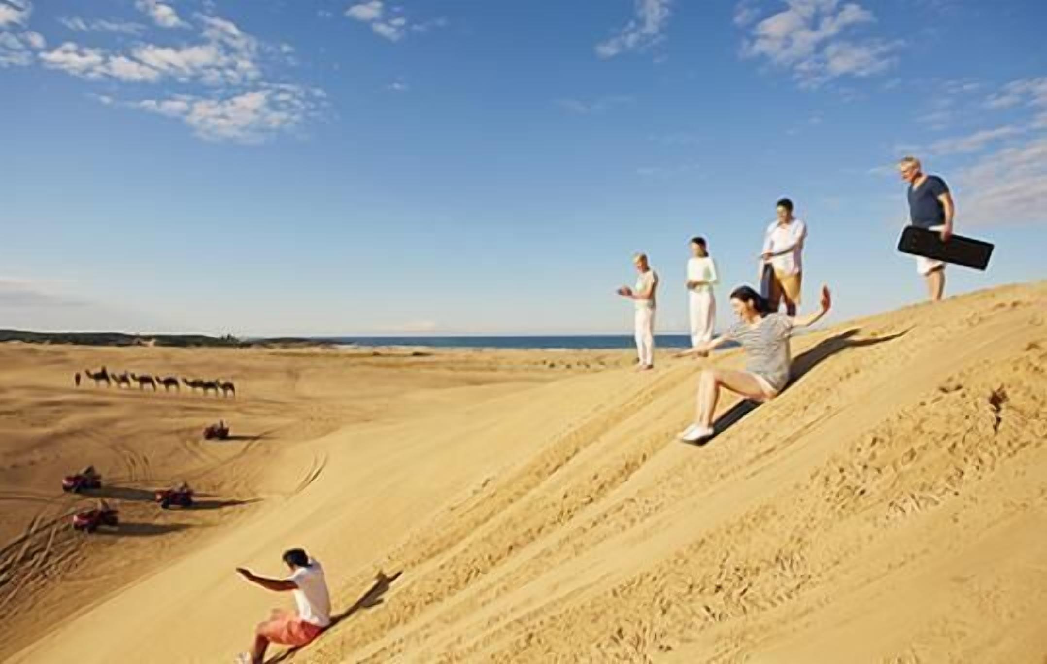 A desert landscape with people on a sand dune, some sitting and others standing, and a camel caravan in the distance.