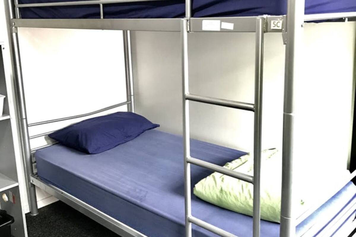 Una cama litera con colchón y almohada azules, barandillas de metal y un ventanal pequeño.