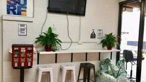 Une pièce équipée d’un téléviseur fixé au mur, d’un distributeur automatique, de deux plantes en pot, d’un fauteuil en forme de sac et d’un mur blanc orné d’un panneau indiquant le mot « SURE ».