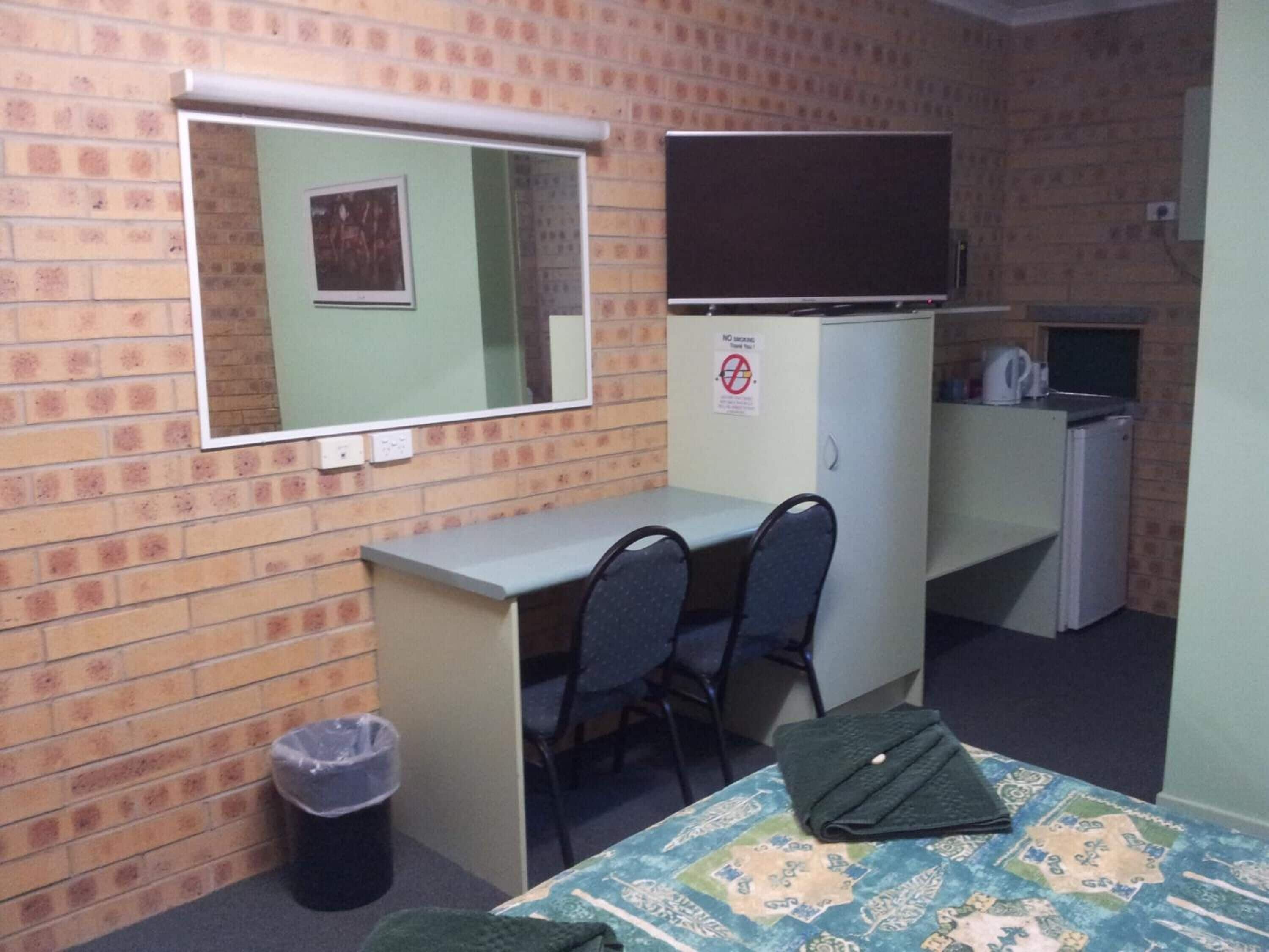 A small, furnished room with a desk, two chairs, a bed, a mirror, a television, and a refrigerator.