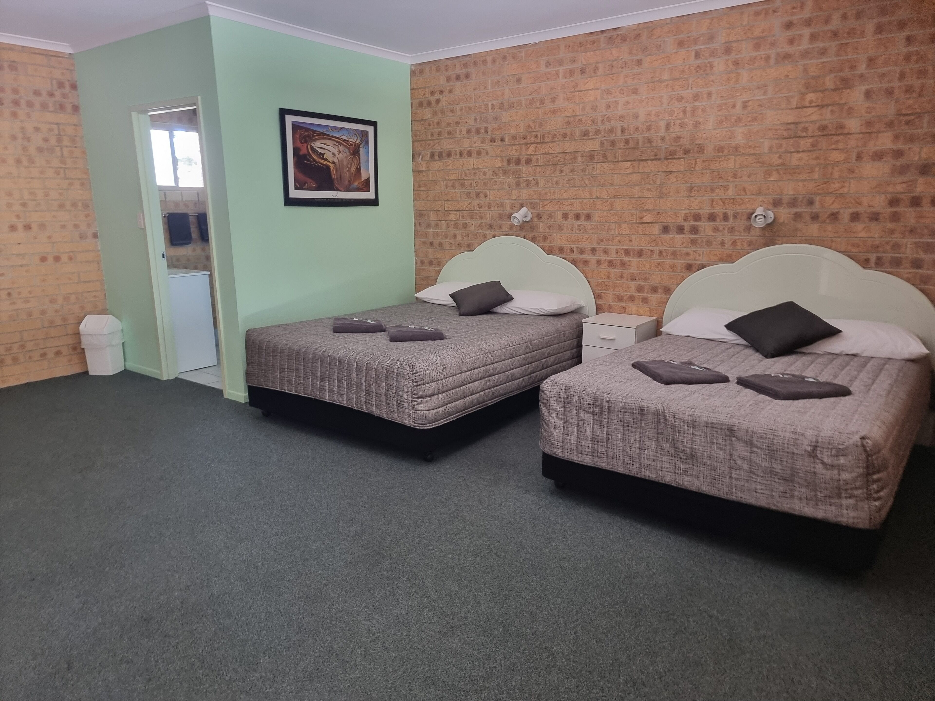 A room with two beds, a small white nightstand, a trash bin, and a framed picture on the wall.