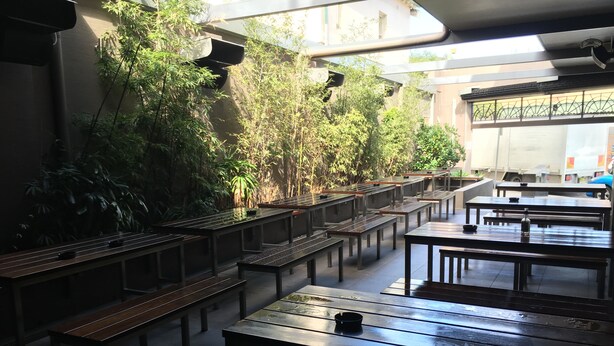 Un amplio espacio interior con mesas y bancos largos, rodeado de abundante vegetación y un techo alto con tragaluces.