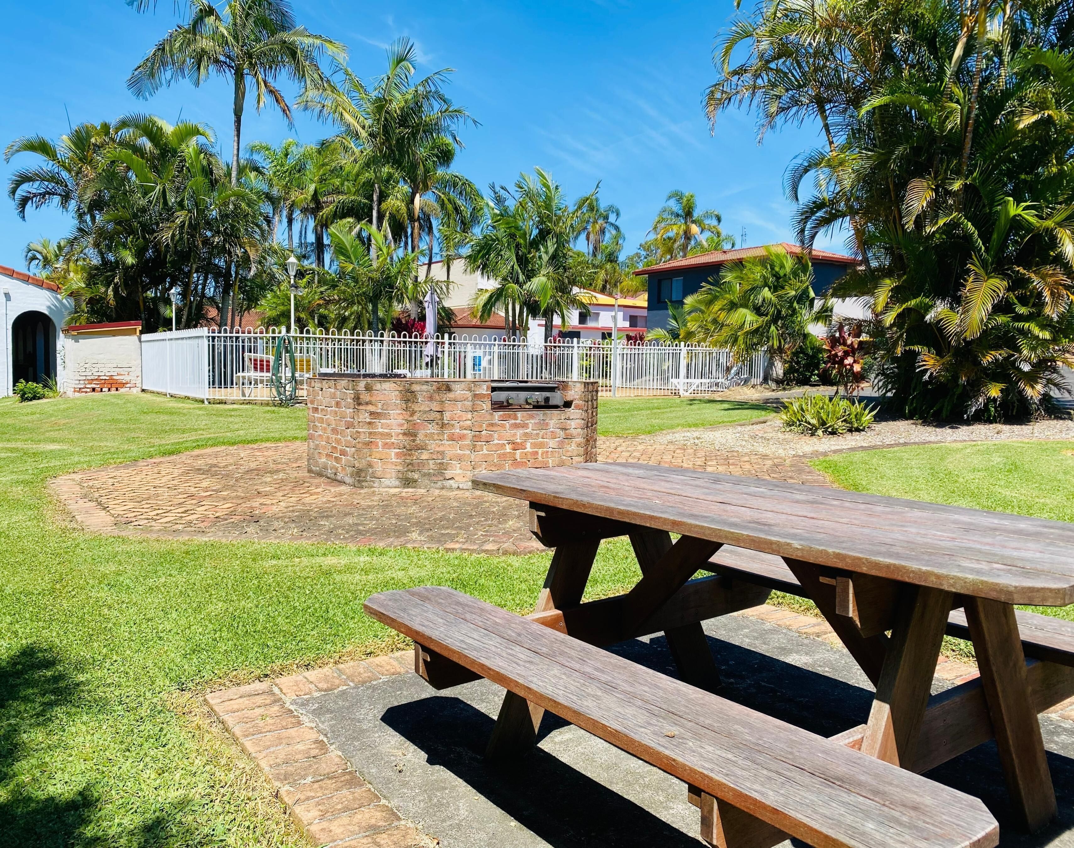 A picnic area with a brick barbecue pit, surrounded by a white fence and palm trees.