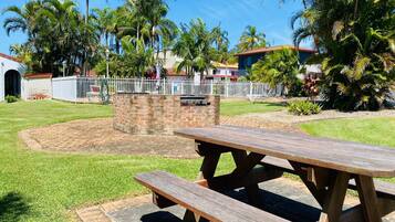 A picnic area with a brick barbecue pit, surrounded by a white fence and palm trees.