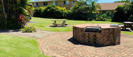 Un patio trasero con un pozo de fuego de ladrillo, mesas de picnic y una casa al fondo.
