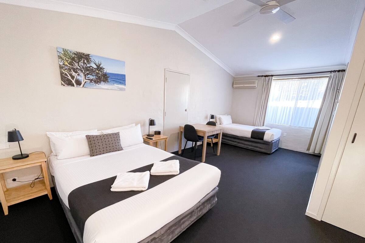 A hotel room with a large bed, a desk, a chair, a small table, a ceiling fan, and a window with curtains.