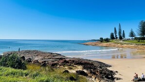 A coastal beach with rocky outcrops, clear blue sky, and a few people enjoying the water. - Costa Rica Motel (South West Rocks)