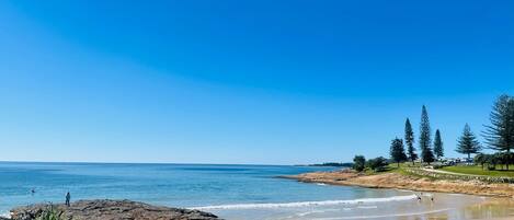 A coastal beach with rocky outcrops, clear blue sky, and a few people enjoying the water.