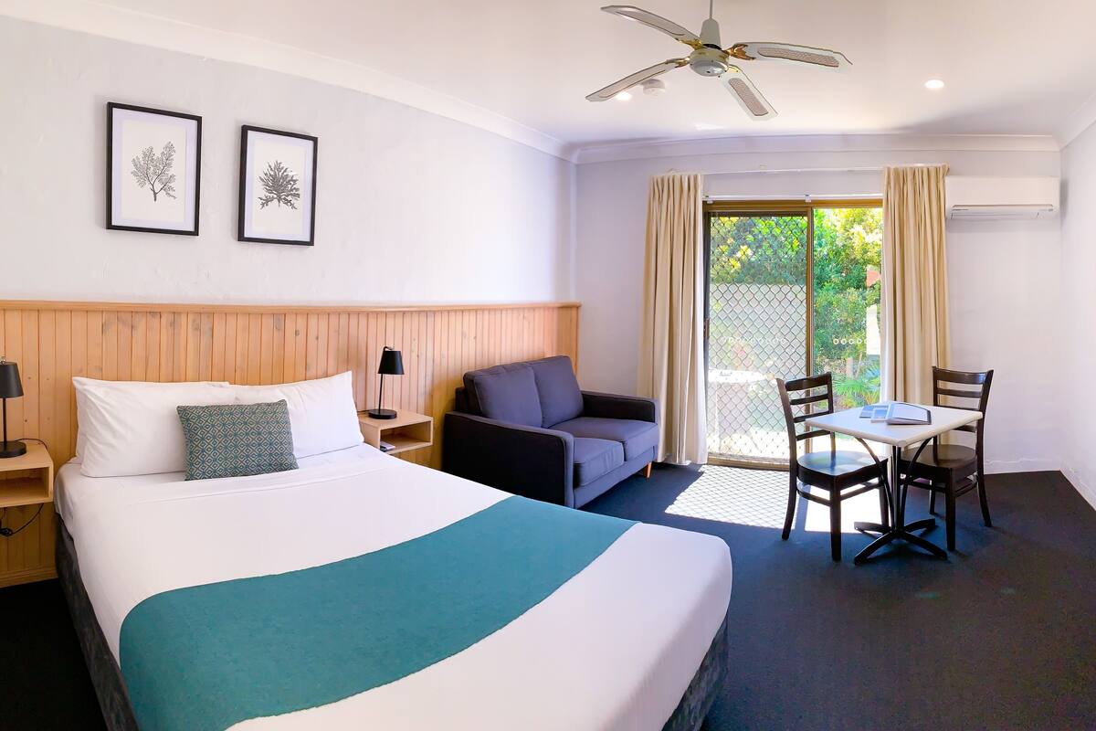 A hotel room with a bed, a sofa, a small table with chairs, a ceiling fan, and a TV mounted on the wall.