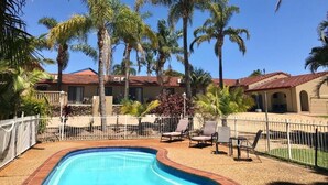 A swimming pool surrounded by palm trees and lounge chairs, with a building in the background. - Costa Rica Motel (South West Rocks)