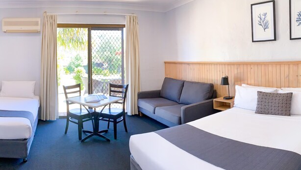 A hotel room with a large bed, a sofa, a small table with chairs, and a desk with a lamp. There is a window with curtains and a view of greenery outside. - Costa Rica Motel (South West Rocks)