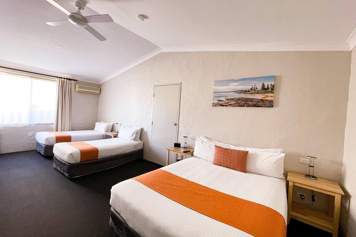 A hotel room with two beds, a ceiling fan, a painting of a beach, and a bedside table with a lamp.