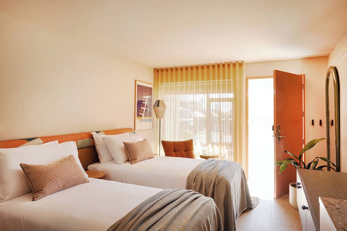 A hotel room with two beds, a bedside table, a lamp, a door, and a potted plant.