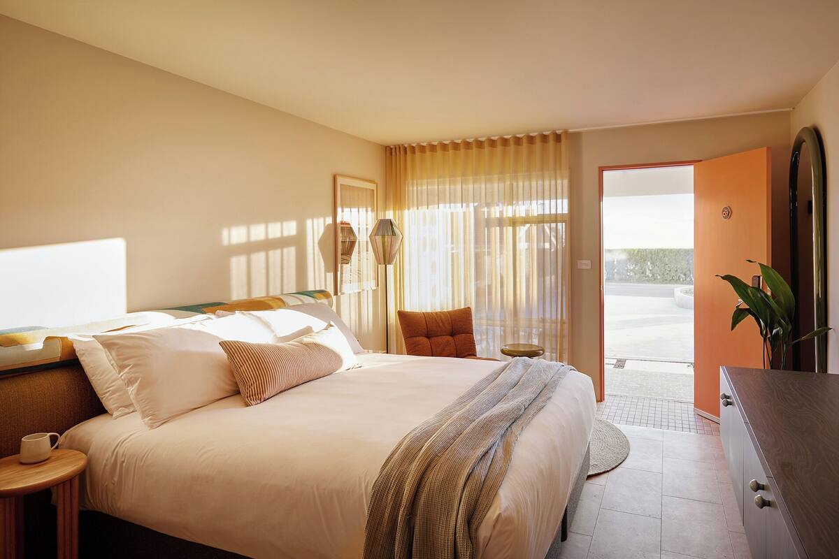 A bedroom with a bed, bedside table, lamp, chair, and a view through a door.