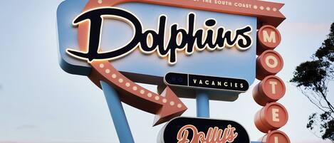 Placa de motel com os textos 'Dolphins' e 'Dolly's Café', um aviso de 'Sem Quartos Disponíveis' e um design retro clássico.