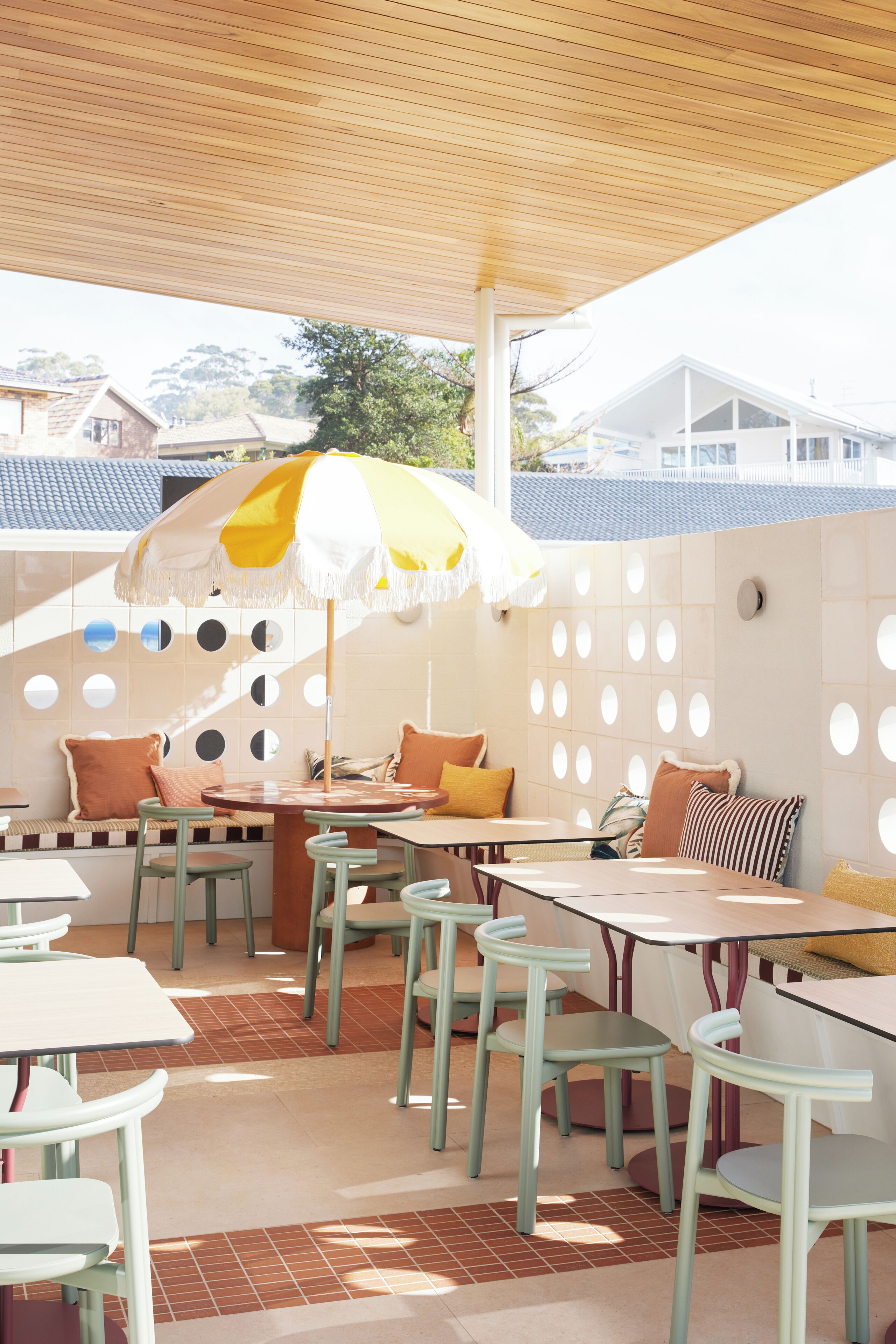 A covered outdoor seating area with tables and chairs, a yellow and white umbrella, and a wall with circular cutouts.
