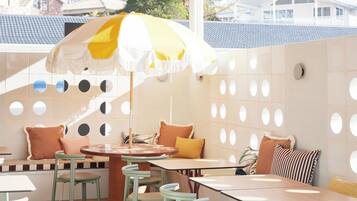 A covered outdoor seating area with tables and chairs, a yellow and white umbrella, and a wall with circular cutouts.