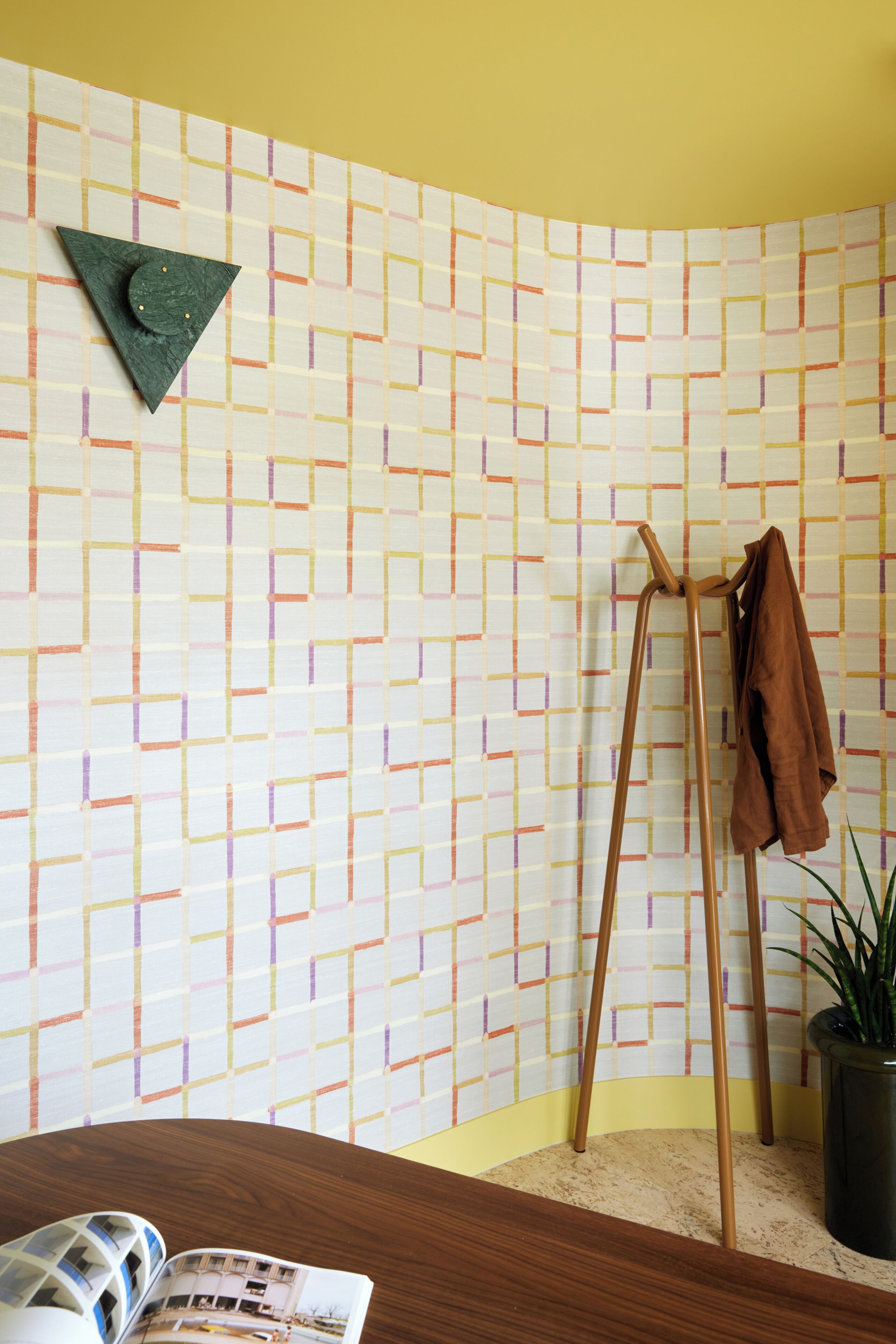 Una habitación con pared estampada, un caballete de madera, una chaqueta marrón, una planta en maceta y una revista sobre una mesa de madera.