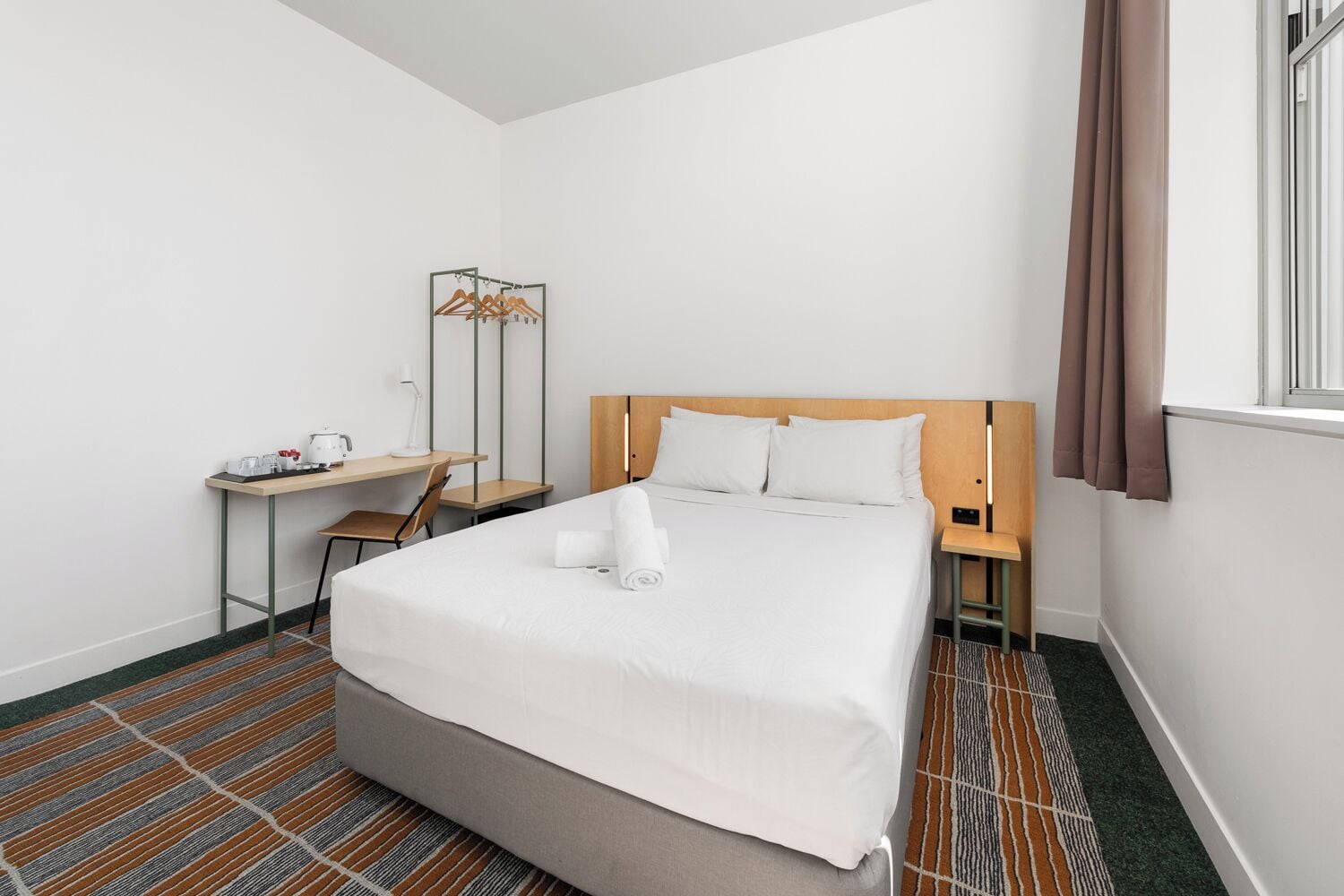A hotel room with a bed, a desk, a chair, a mirror, and a window with curtains.