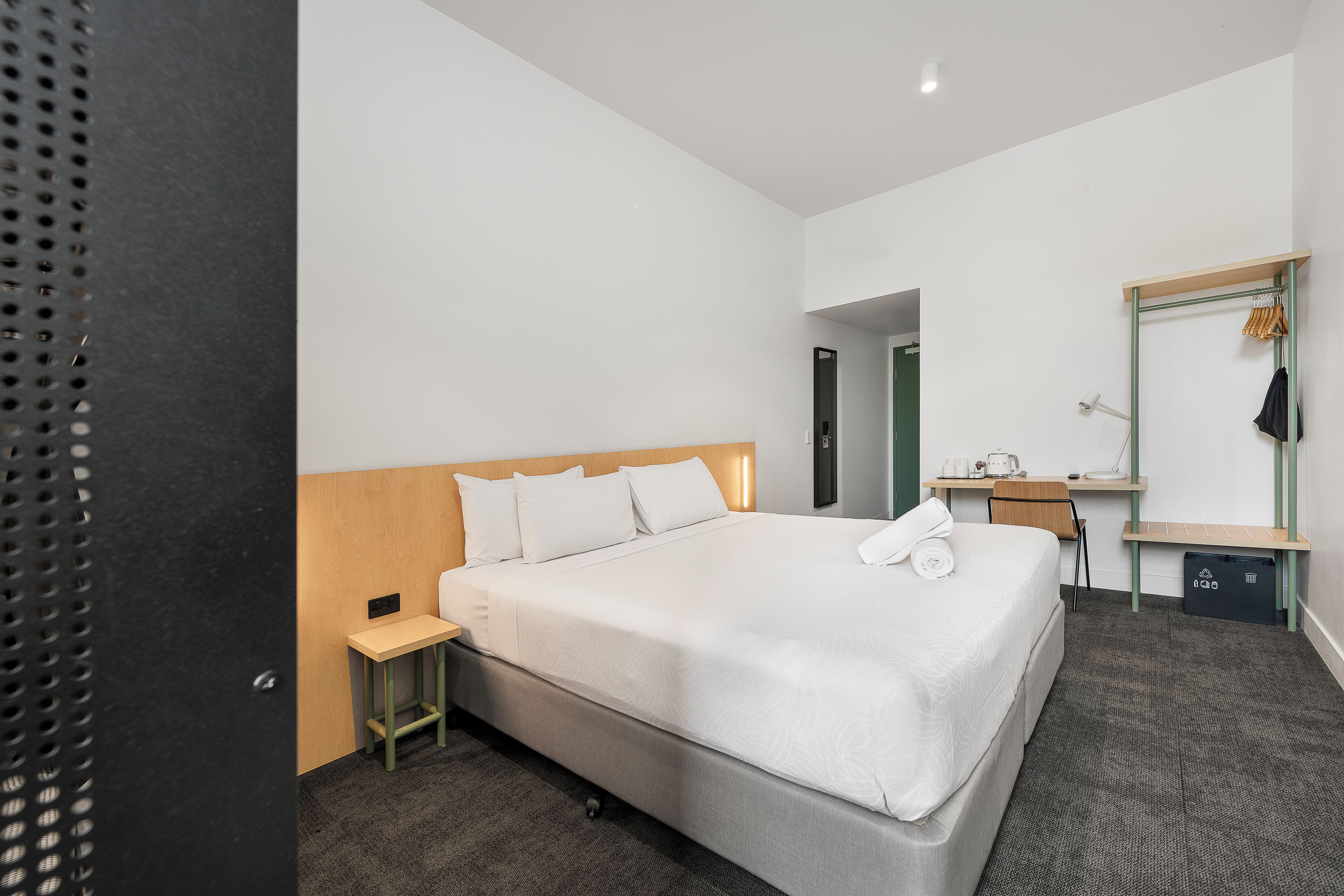 A modern hotel room with a large bed, a small desk, a mirror, and a wardrobe.