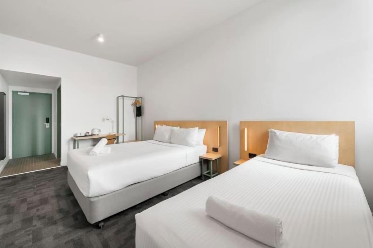 A hotel room with two beds, a small bedside table with a lamp, a mirror, and a green door.