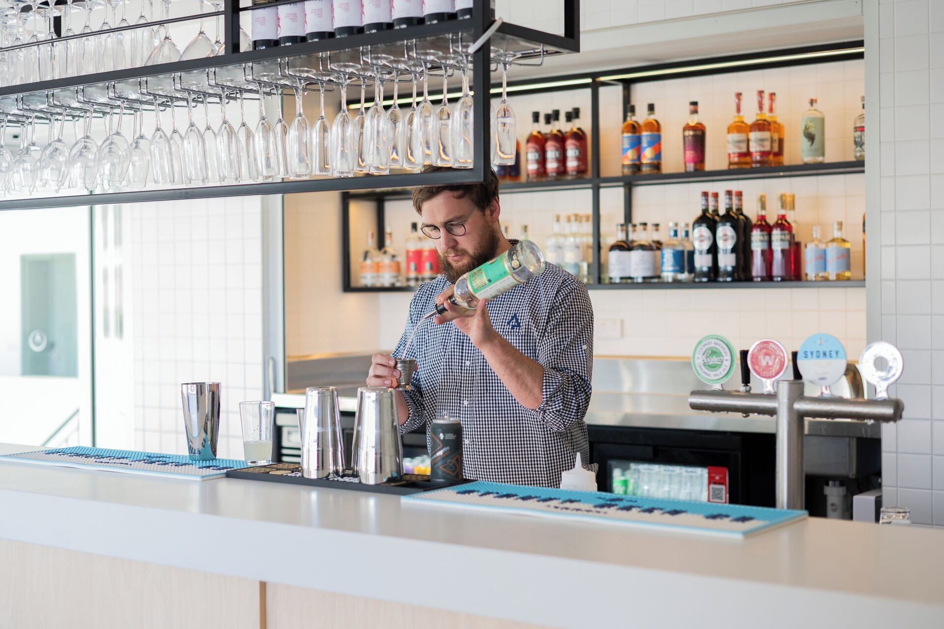 A bar area with a bartender holding a bottle, glasses on a rack, and shelves with bottles.