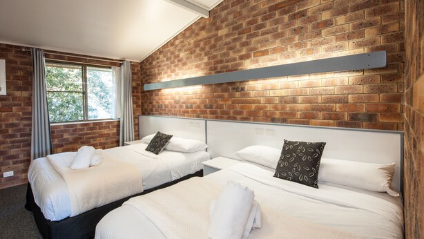 A hotel room with two beds, a brick wall, a window with curtains, and a metal shelf above the beds. - Allan Cunningham Motel (Kearneys Spring)