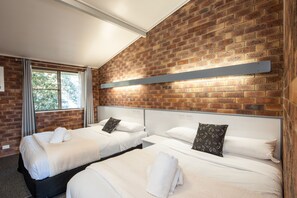 A hotel room with two beds, a brick wall, a window with curtains, and a metal shelf above the beds. - Allan Cunningham Motel (Kearneys Spring)