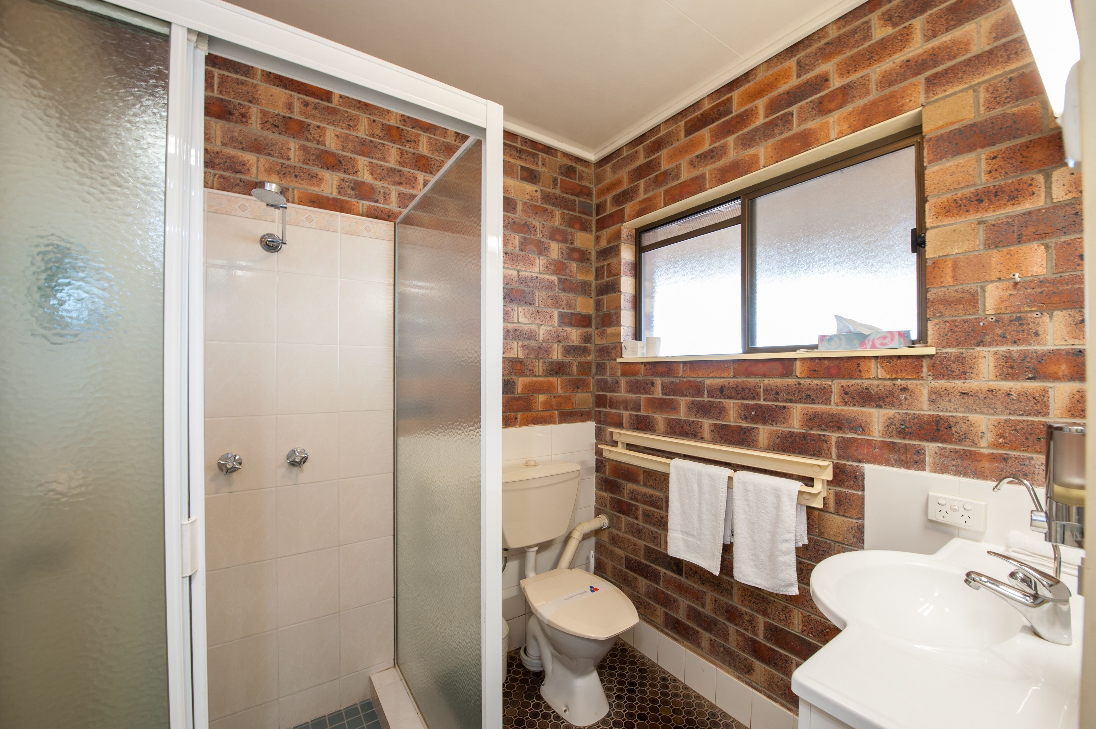 A bathroom with a shower, toilet, and sink, featuring a brick wall and a window.