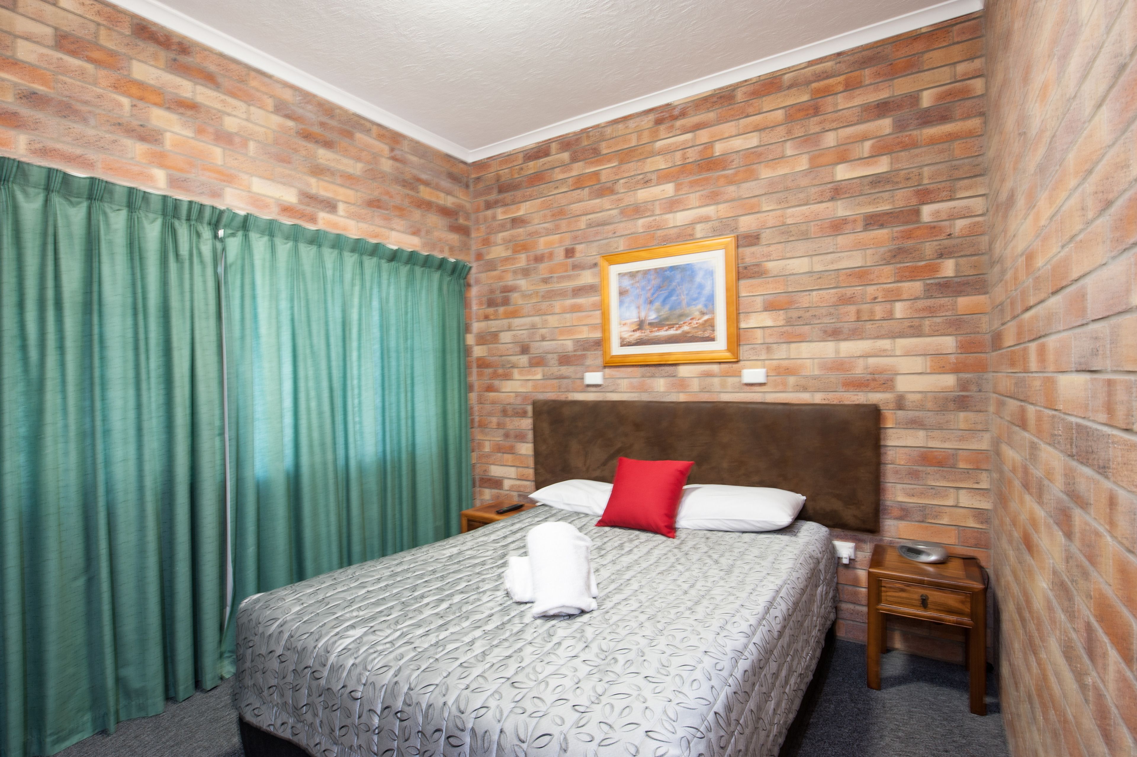A hotel room with a brick-patterned wall, a bed with a patterned bedspread, a red pillow, and a framed picture above the bed.