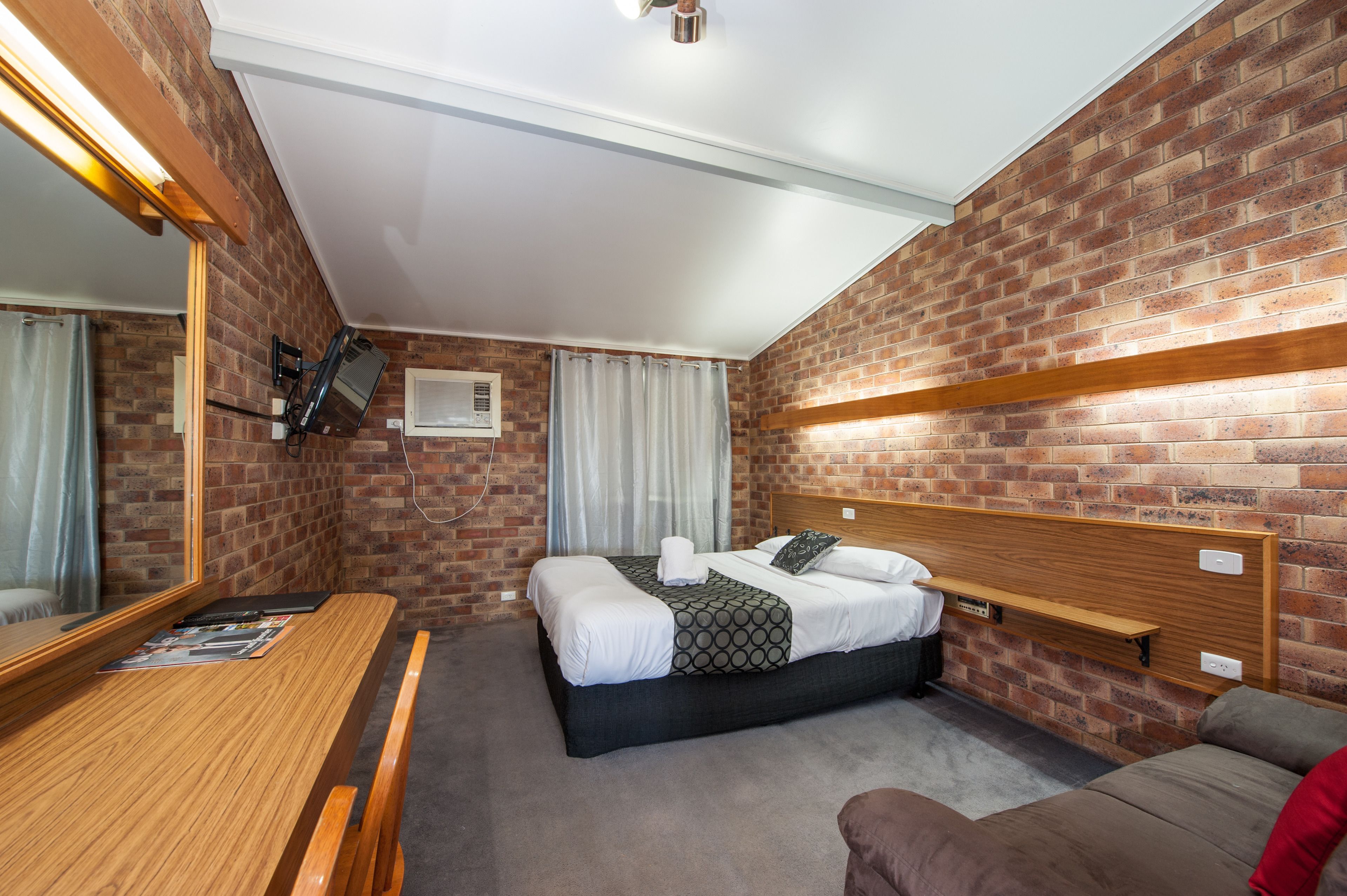 A hotel room with a brick wall, a wooden headboard, a bed with white linens, a wooden desk, and a brown sofa.