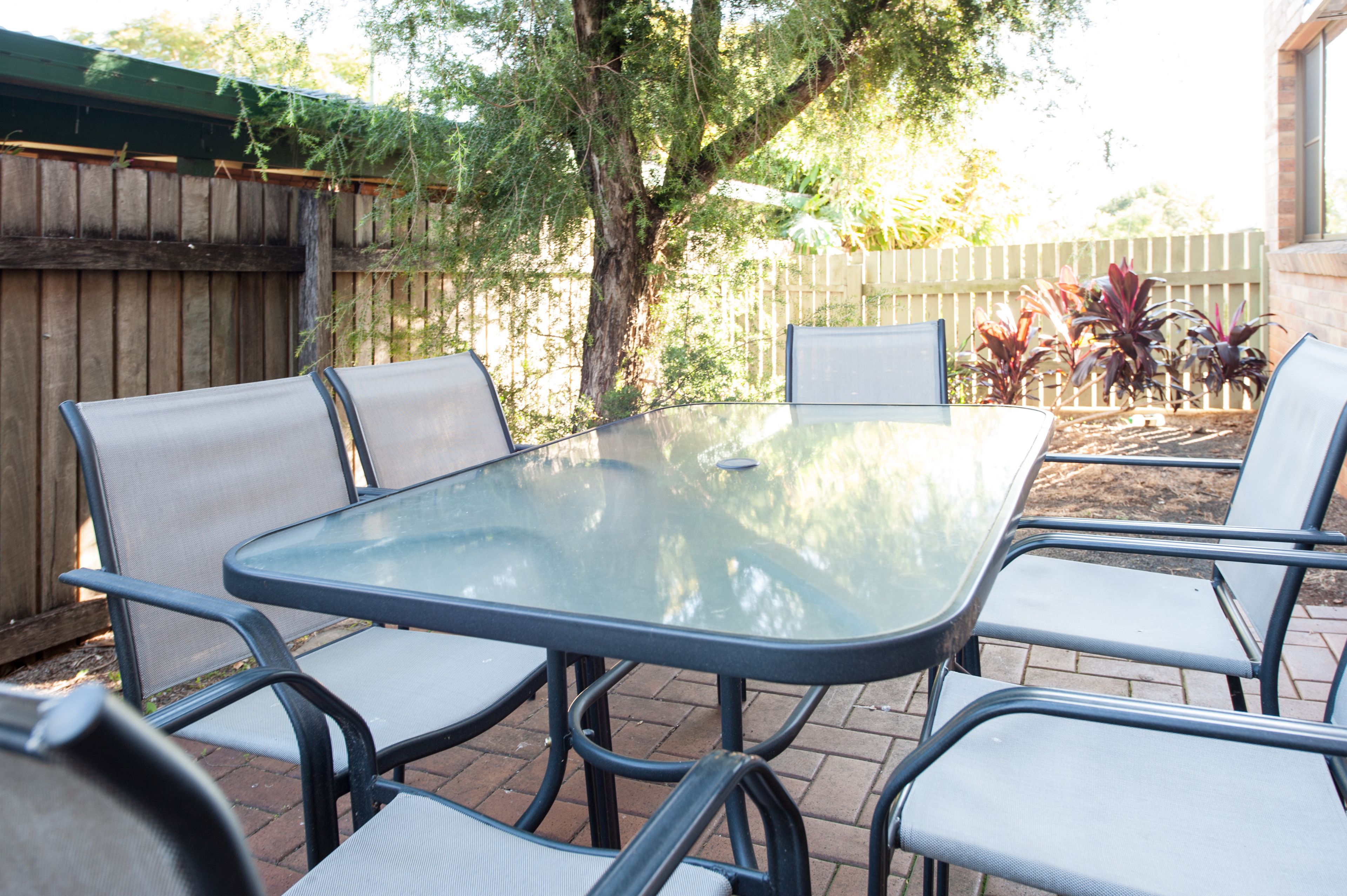 Een terras met een glazen tafel en metalen stoelen, omringd door een houten hek en beplanting.