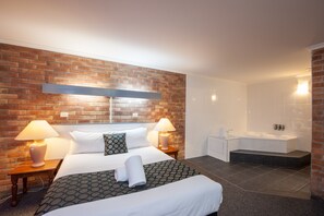 Una habitación de hotel con pared de ladrillo, una cama grande con ropa de cama estampada, dos lámparas de noche, una mesita de noche, un baño con bañera y zona de ducha.