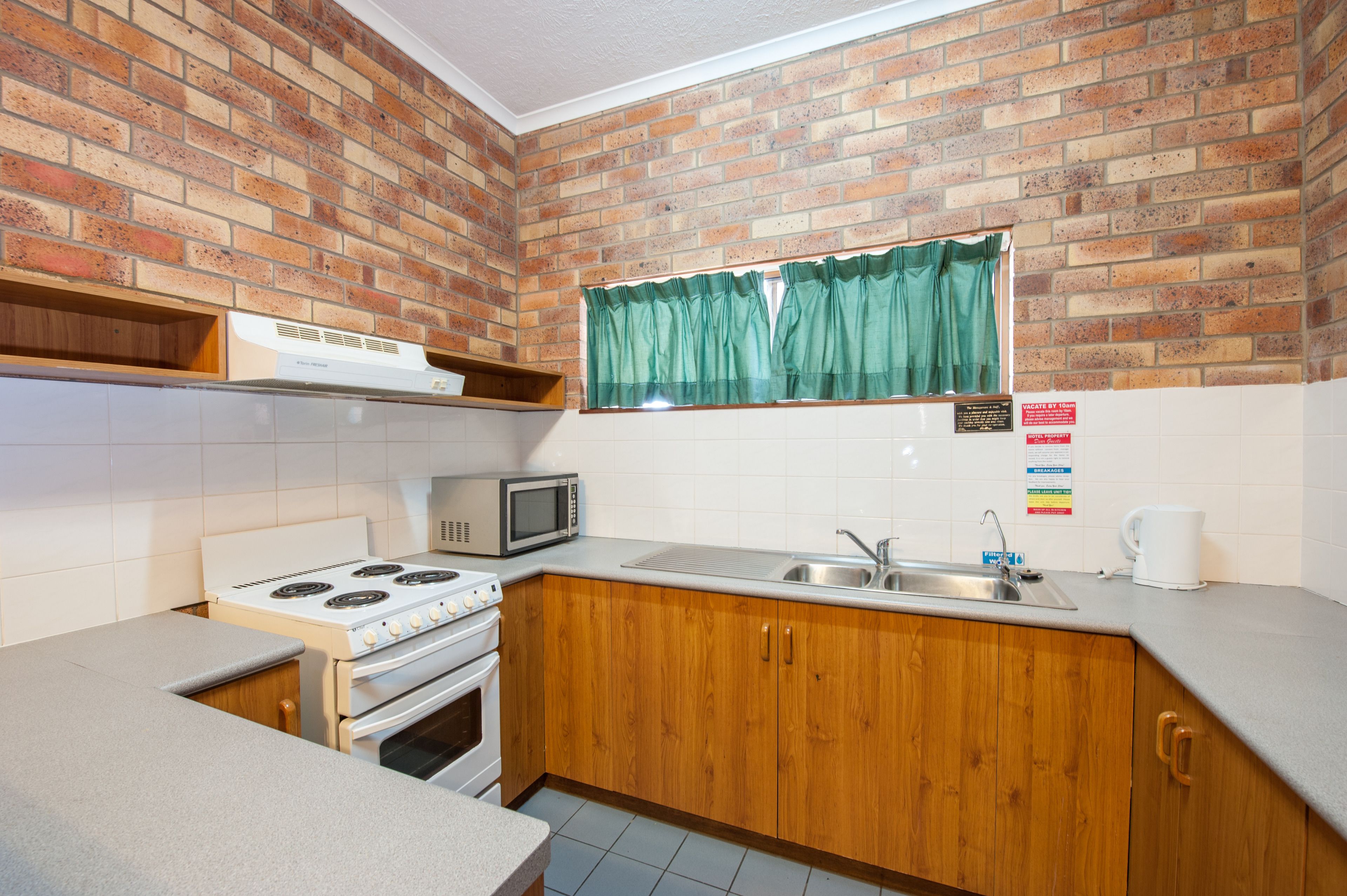A kitchen with a brick wall, wooden cabinets, a microwave, a stove, and a window with green curtains.