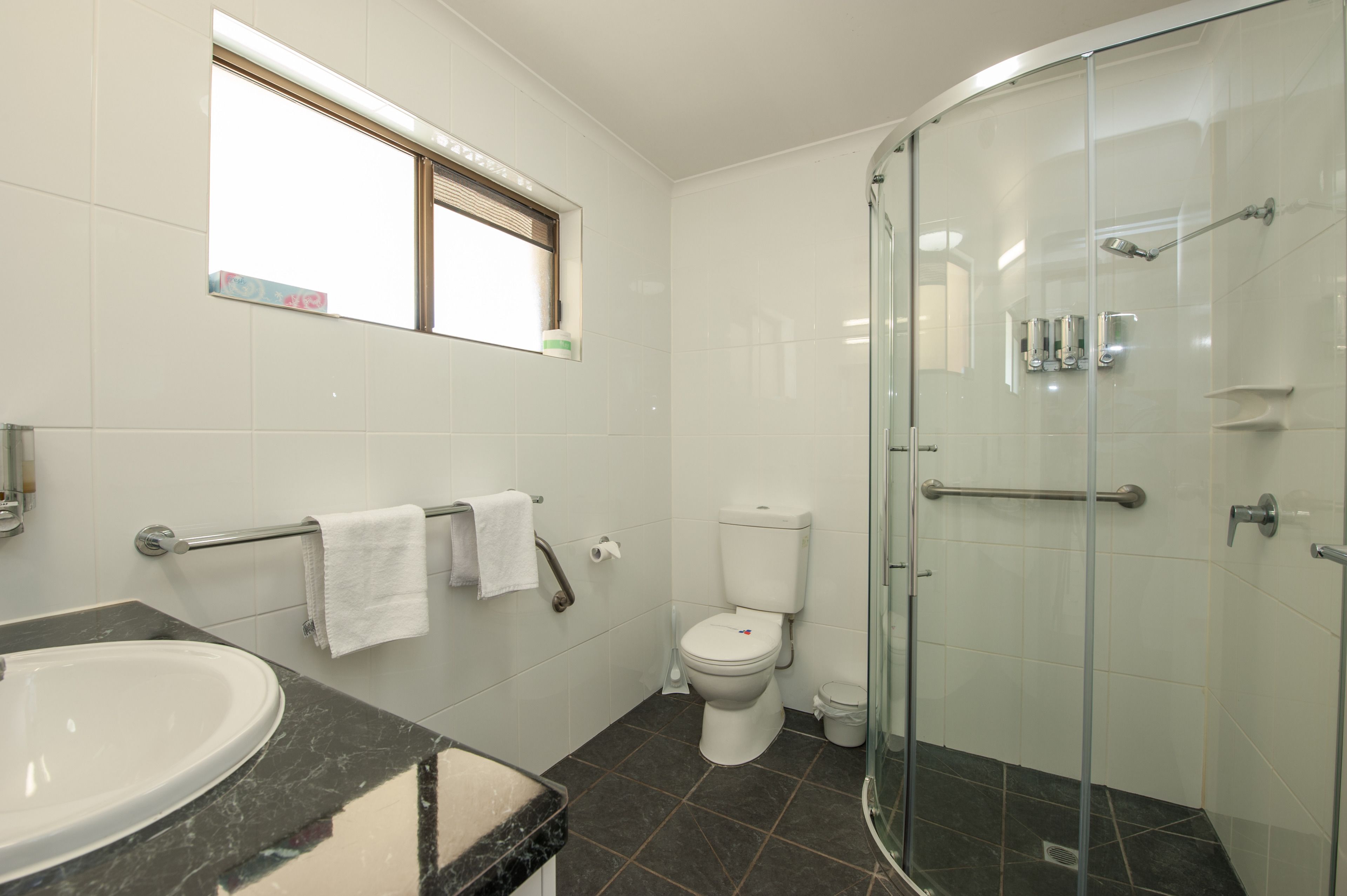 Ein Badezimmer mit Duschkabine aus Glas, Toilette, Waschbecken mit Marmorplatte und einem Fenster mit Jalousien.