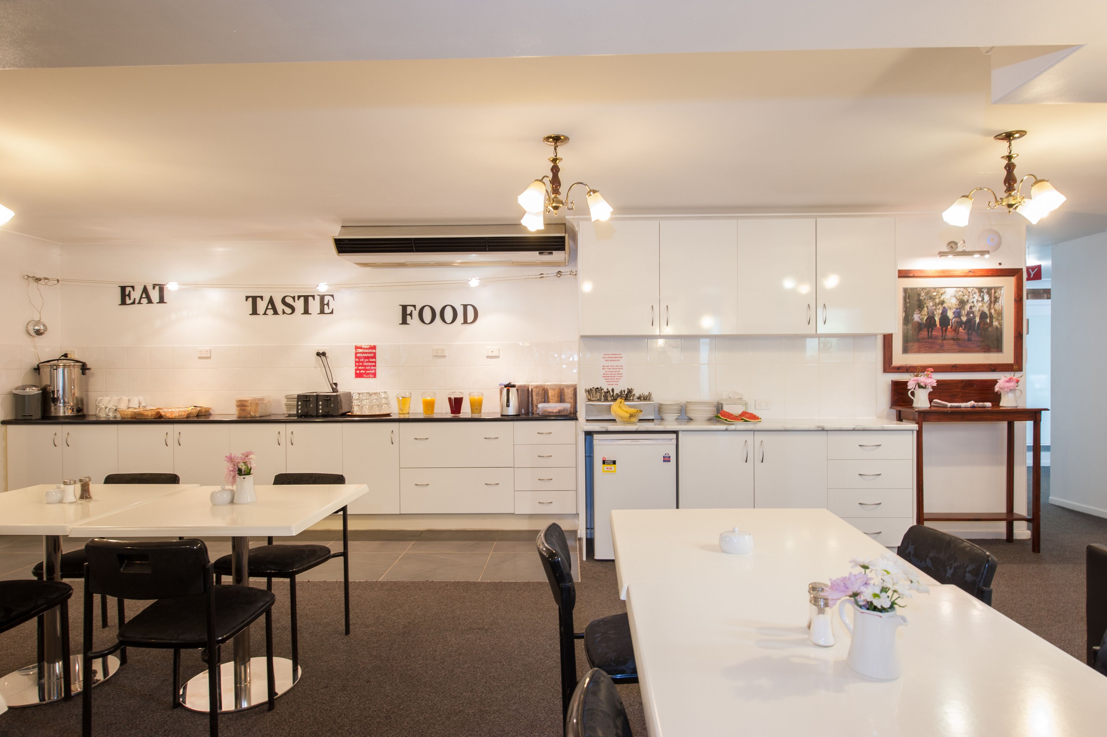 Een moderne keuken met witte kasten, een wit aanrechtblad en een eethoek met een witte tafel en stoelen. Er staan decoratieve items op de tafel, en een bord op de muur met de tekst 'EAT TASTE FOOD'.
