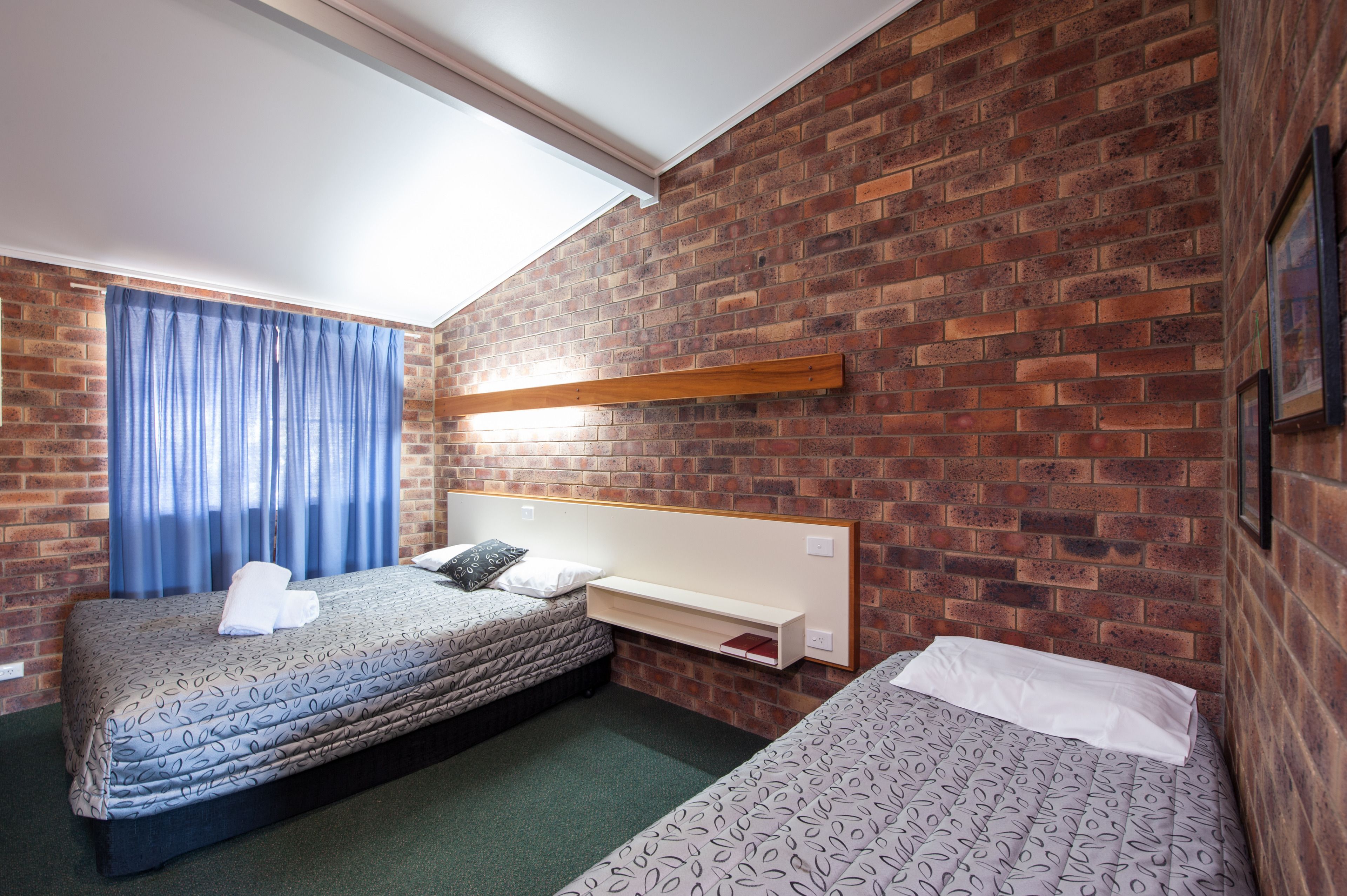 A hotel room with two beds, a brick wall, a window with blue curtains, and a wooden shelf above the beds.