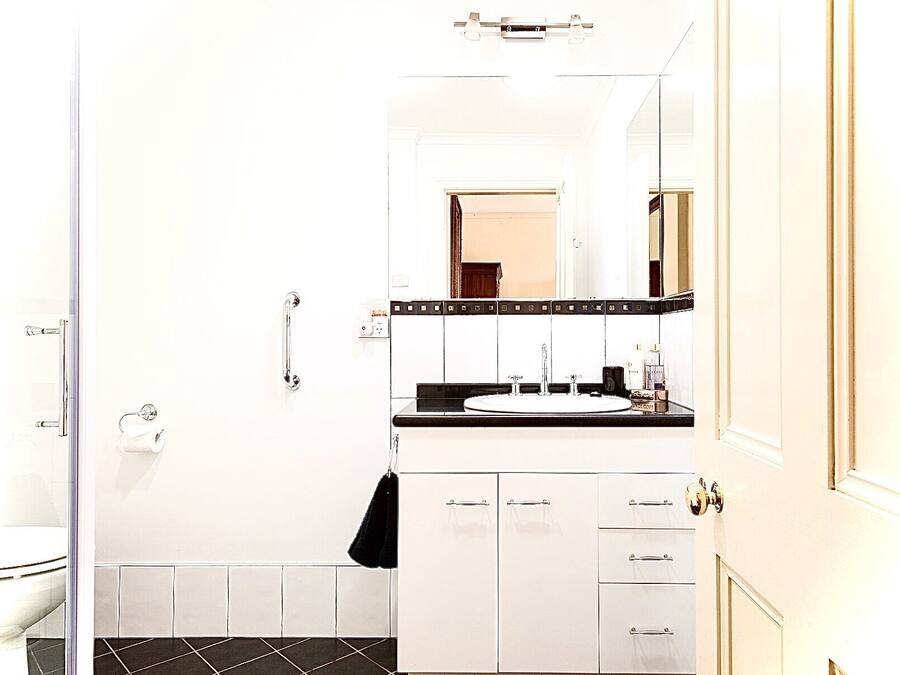 Een badkamer met een douchecabine van glas, een witte wastafel met een zwart aanrechtblad, en een spiegel boven de wastafel.