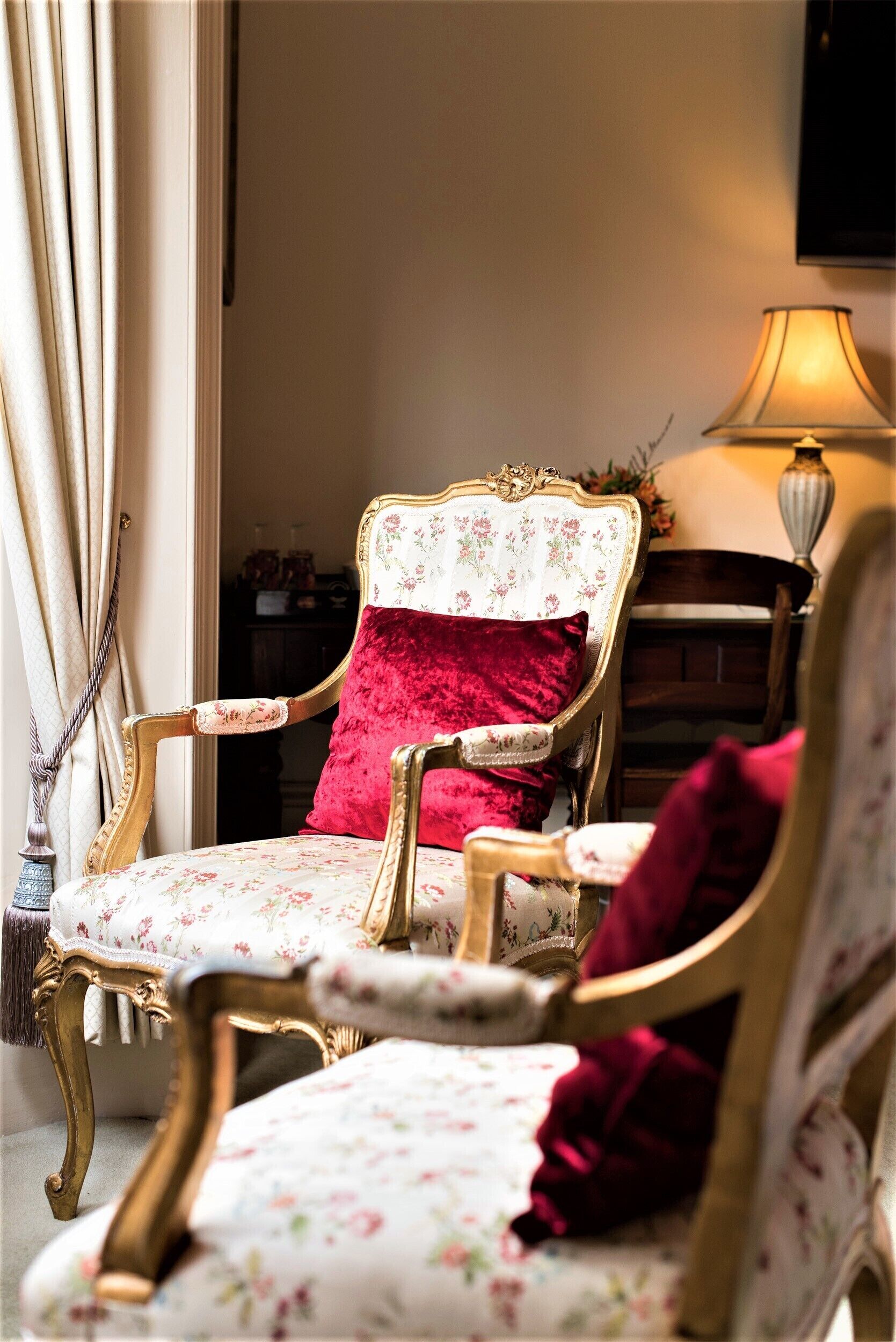 A room with a vintage-style chair with floral upholstery and a red cushion, a lamp, and a curtain.