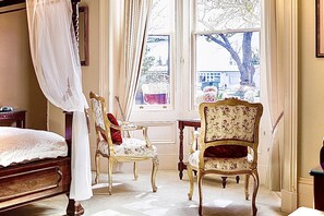 A bedroom with a large bed, two ornate chairs, a small table, and a window with a view of trees and buildings. - Kurrajong House Launceston (East Launceston)