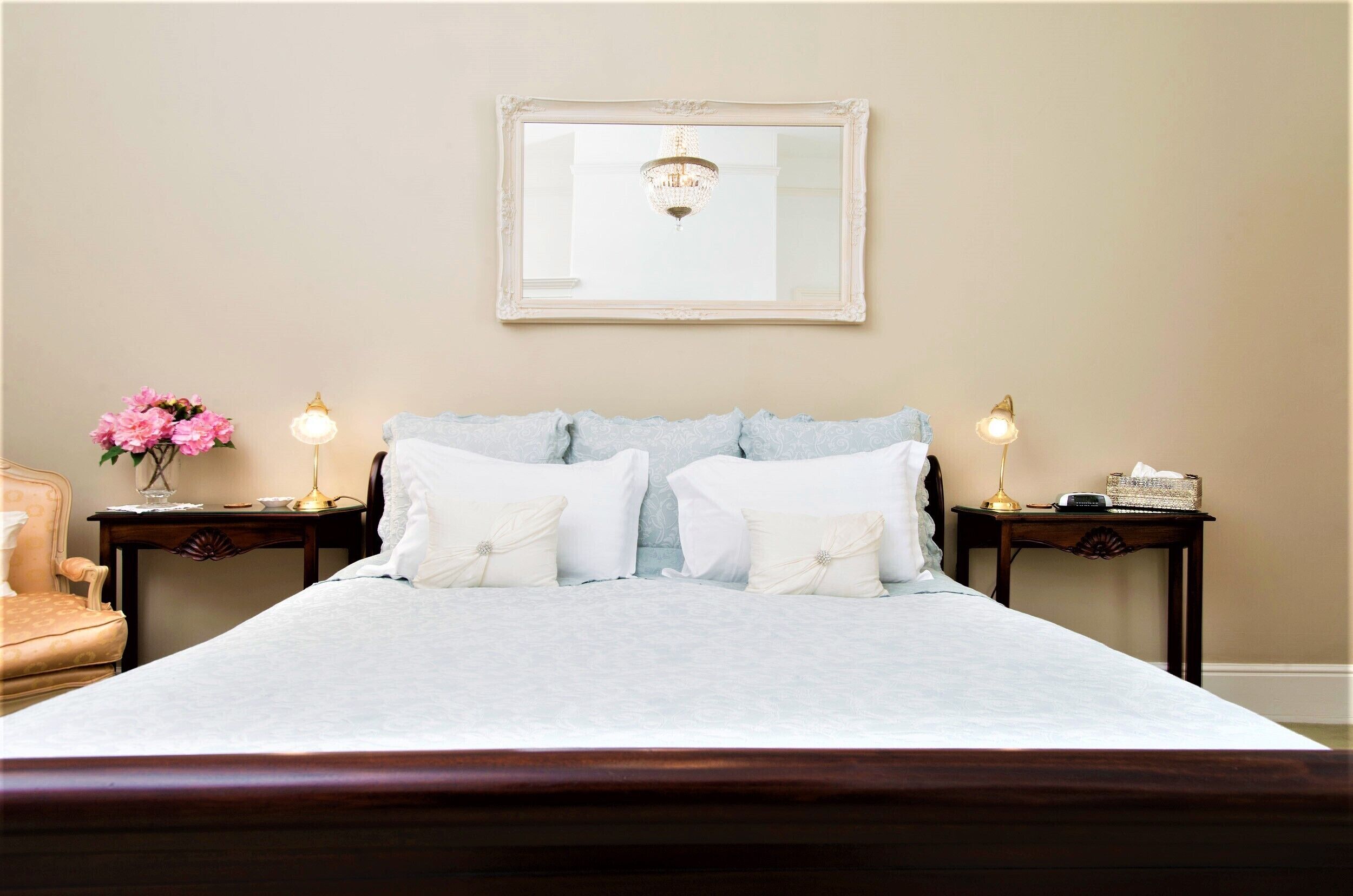 A neatly made bed with white linens, two bedside tables with lamps, a vase of pink flowers, and a mirror on the wall.