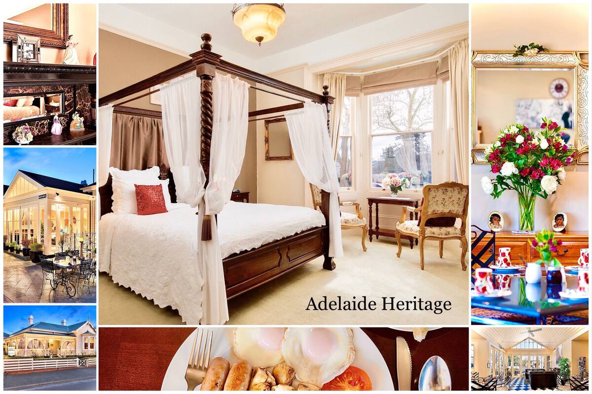 A four-poster bed with white linens, a dining area with a table set for two, and a view of the Adelaide Heritage building.