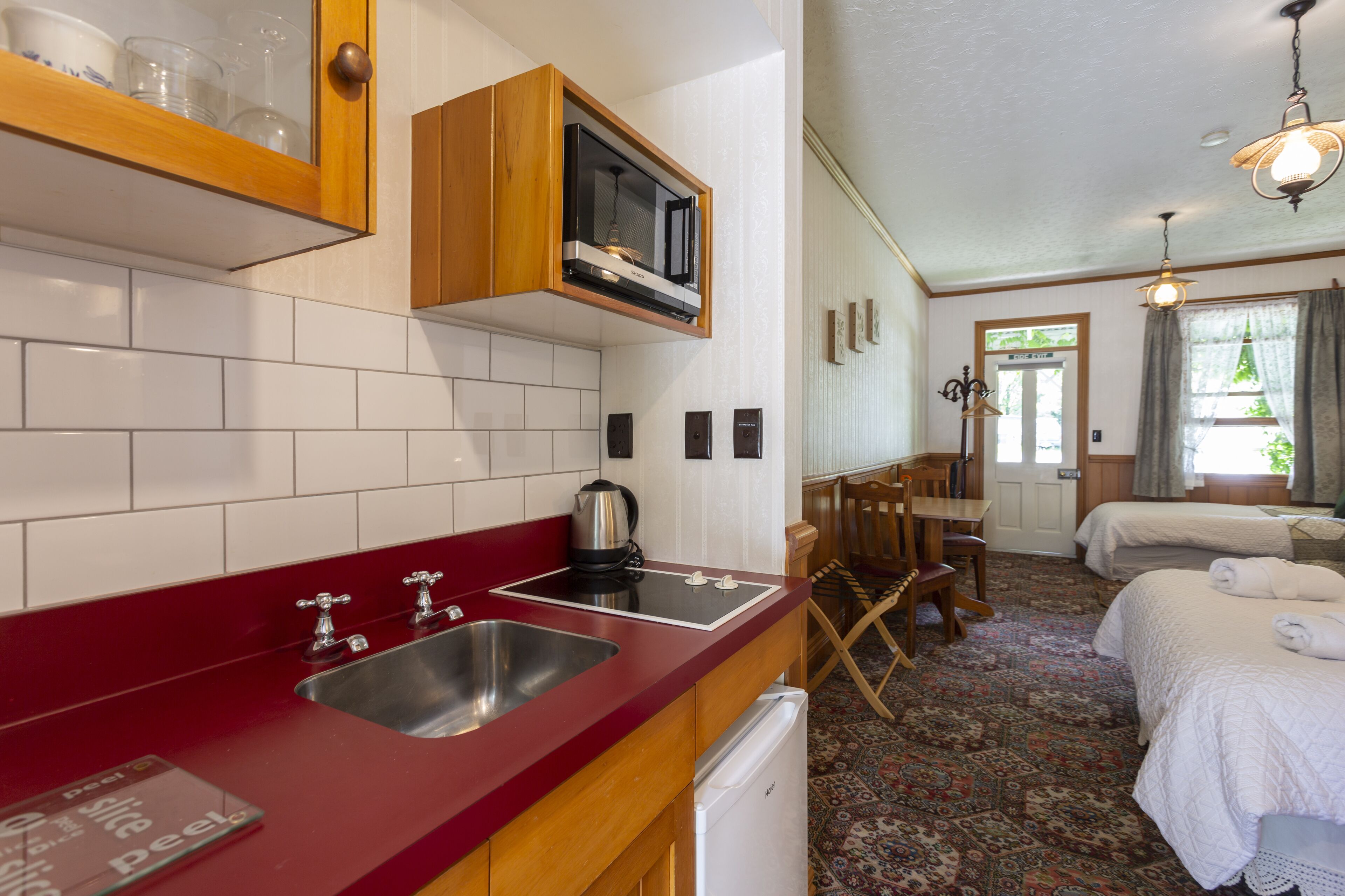 Una cocina con encimera roja, pileta de acero inoxidable y armarios de madera. Hay un microondas sobre la encimera y un refrigerador debajo. La habitación también incluye una zona de comedor con mesa y sillas, y un dormitorio con cama y ventana.
