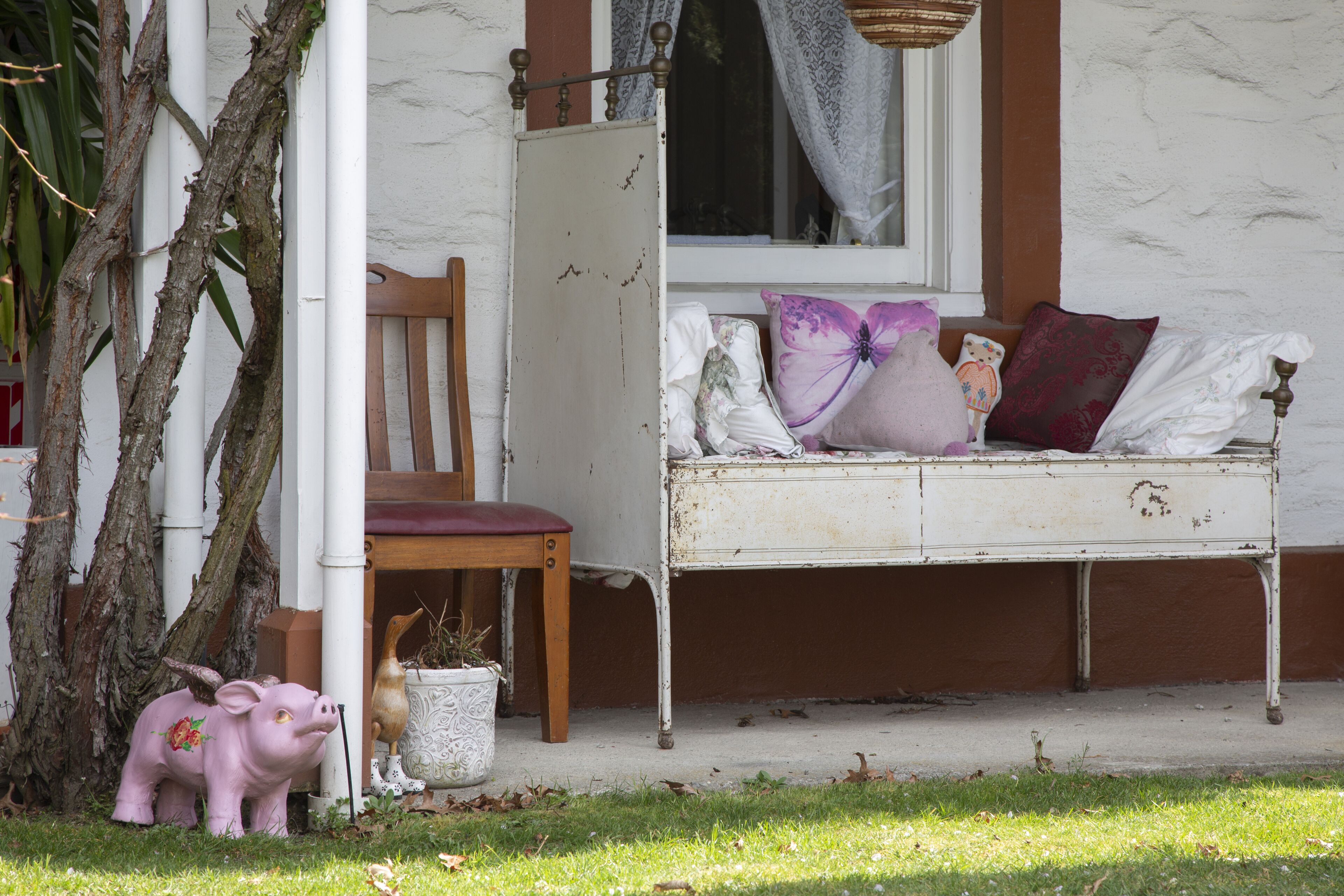 A rustic outdoor seating area with a white metal bench, a wooden chair, and a pink pig sculpture.