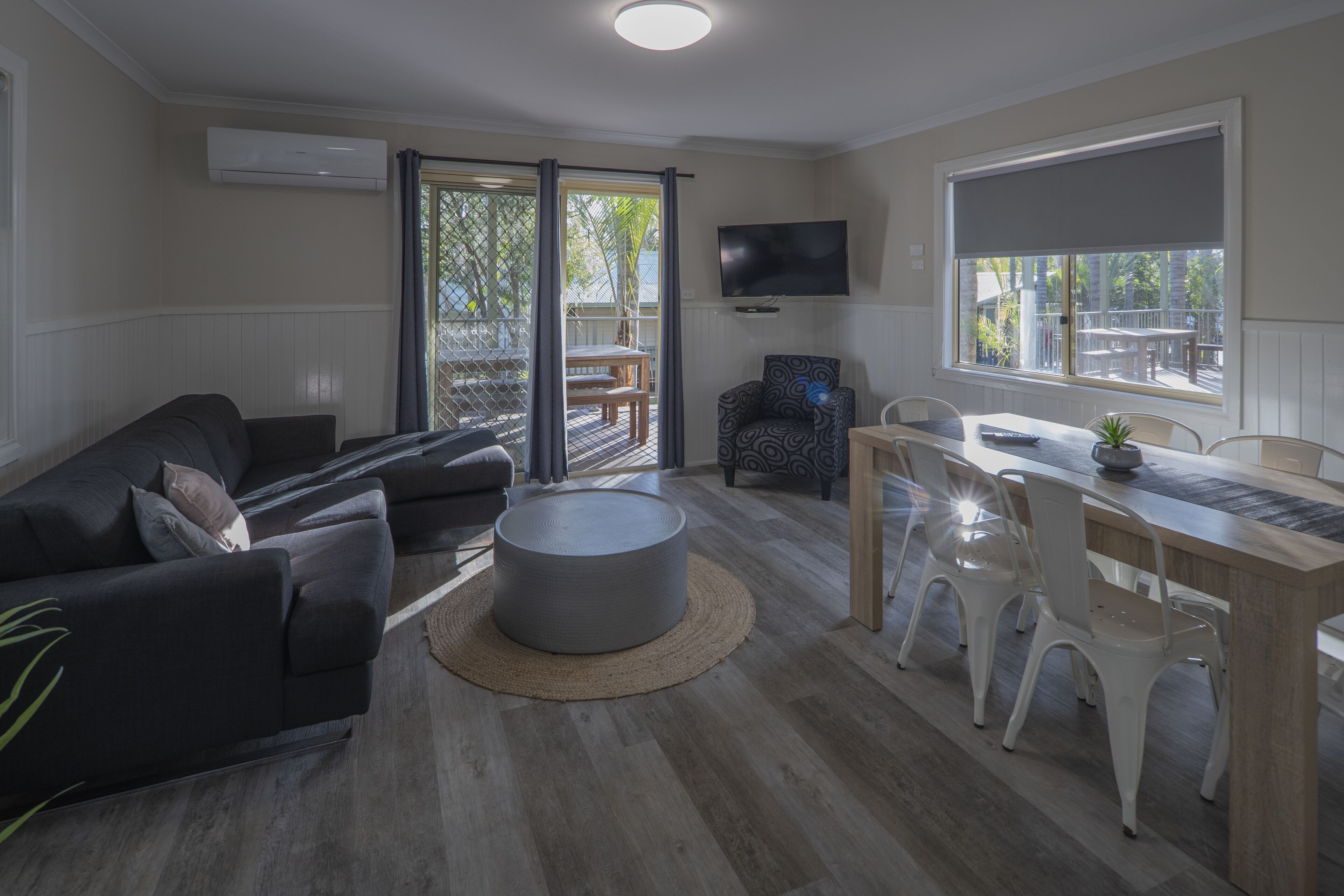 A modern living room with a sofa, ottoman, dining table, and chairs. There is a TV mounted on the wall and a window with a view of the outdoors.