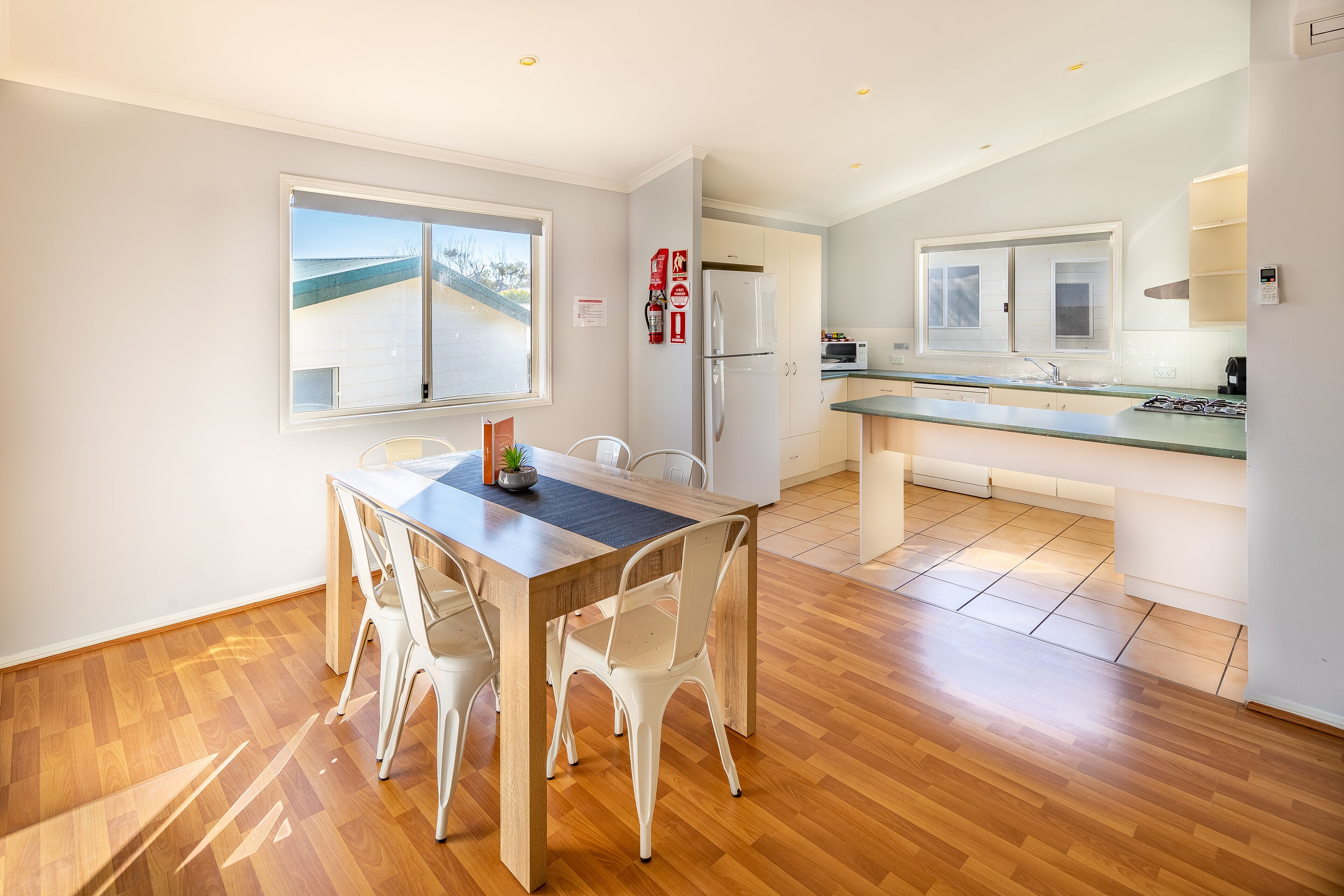 A modern kitchen with a wooden dining table and chairs, a refrigerator, and a view of the beach through a window.