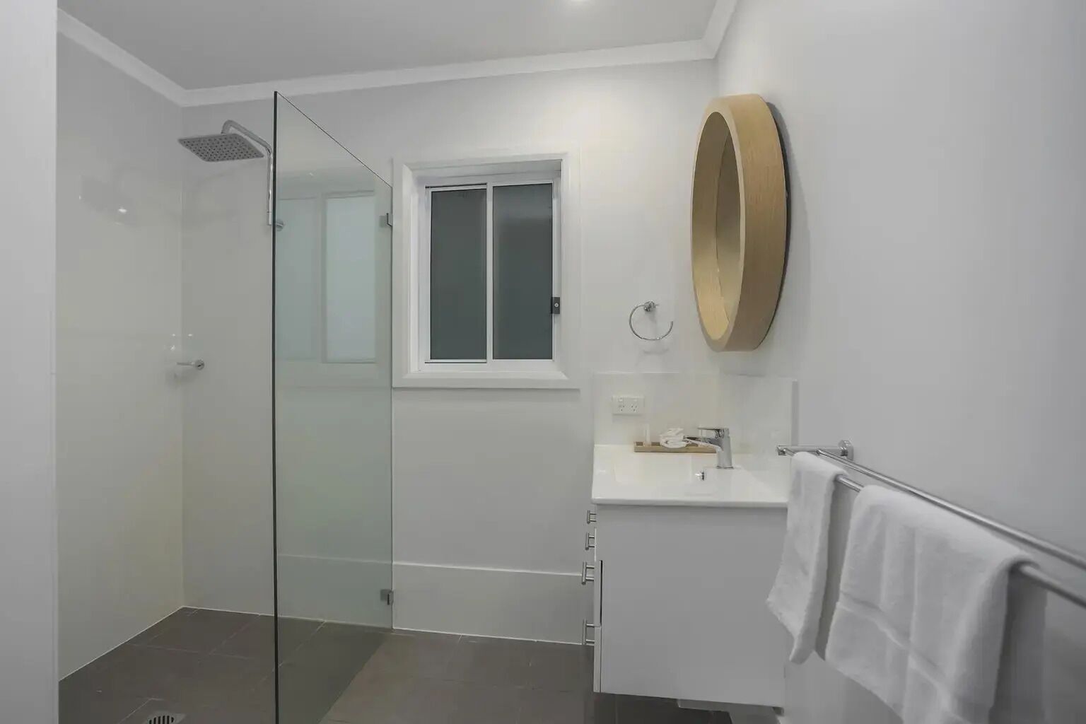 A modern bathroom with a glass shower enclosure, a white sink with a gold mirror, and a window above the sink.