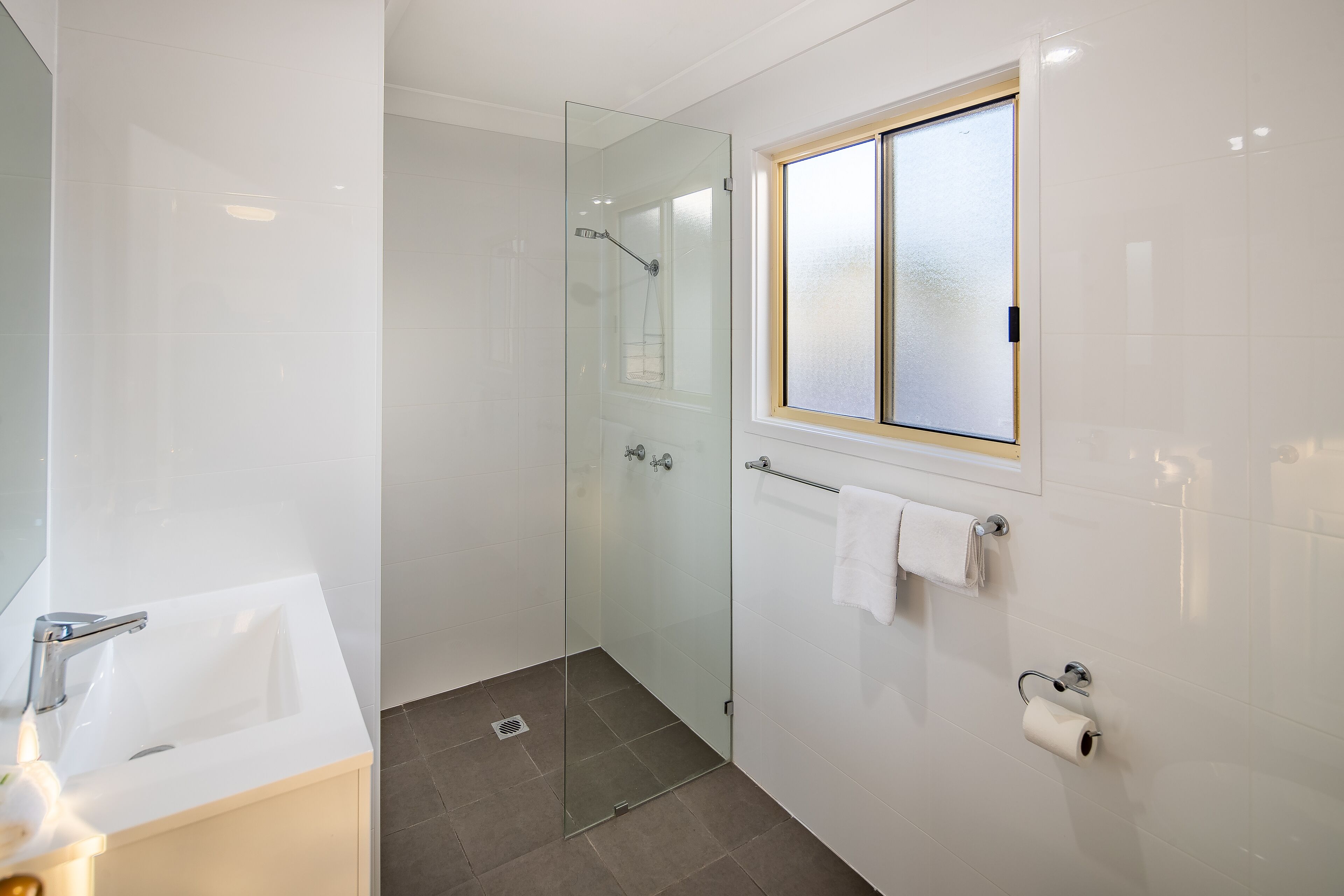 A modern bathroom with a glass-enclosed shower, a white sink, and a window above the toilet.