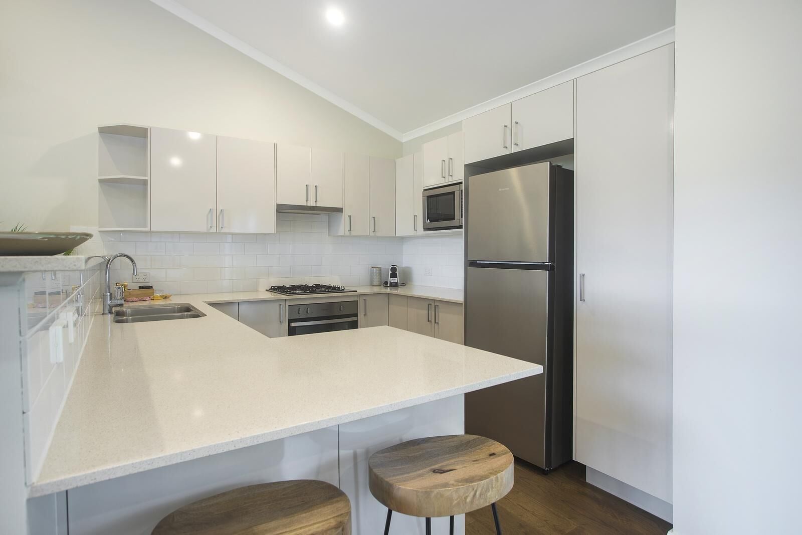 A modern kitchen with white cabinetry, a stainless steel refrigerator, a microwave, and a gas stove. There are two wooden stools at the counter.