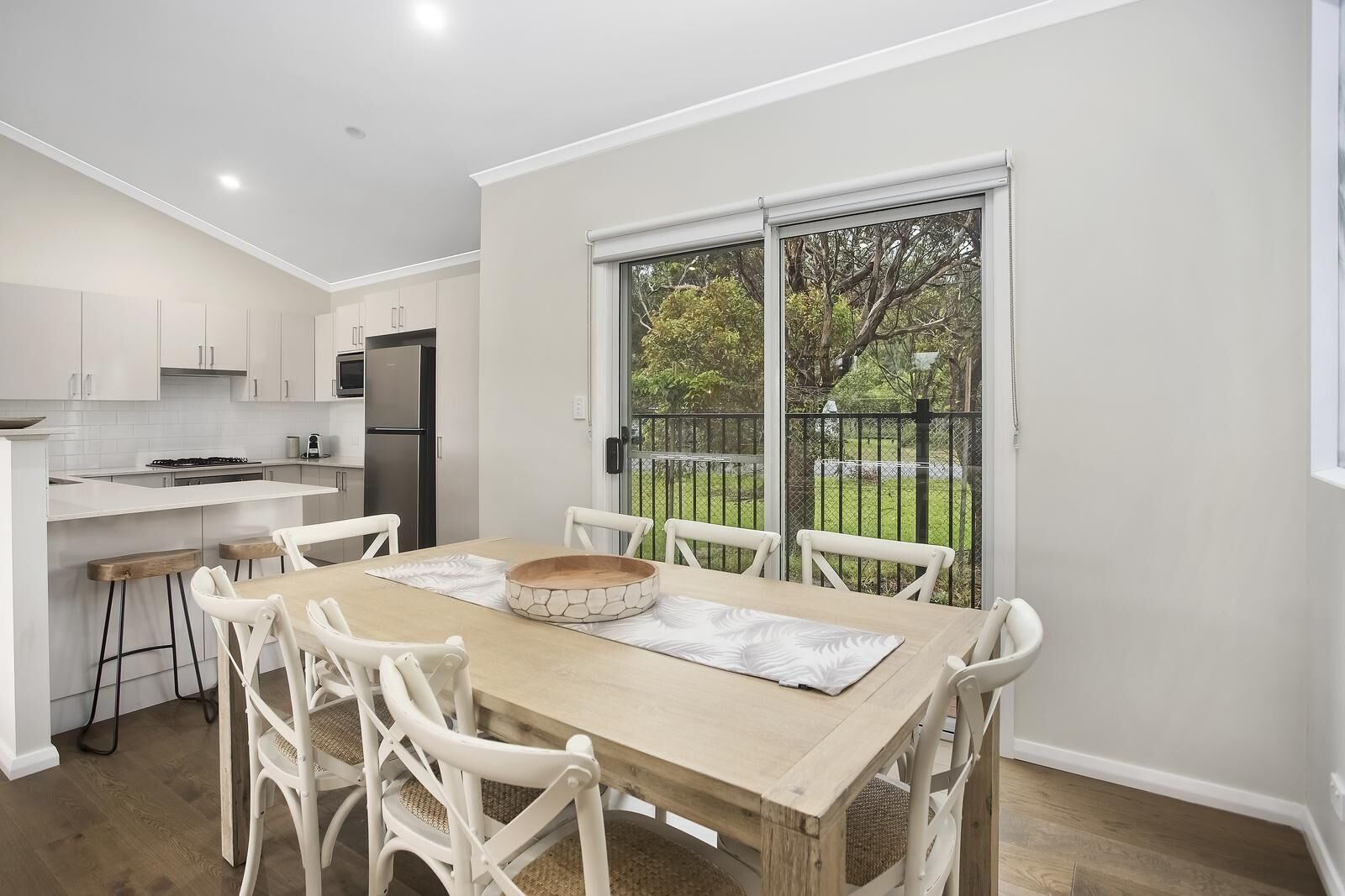 A modern kitchen with white cabinets, a dining table with chairs, and a view of a fenced garden through a sliding glass door.